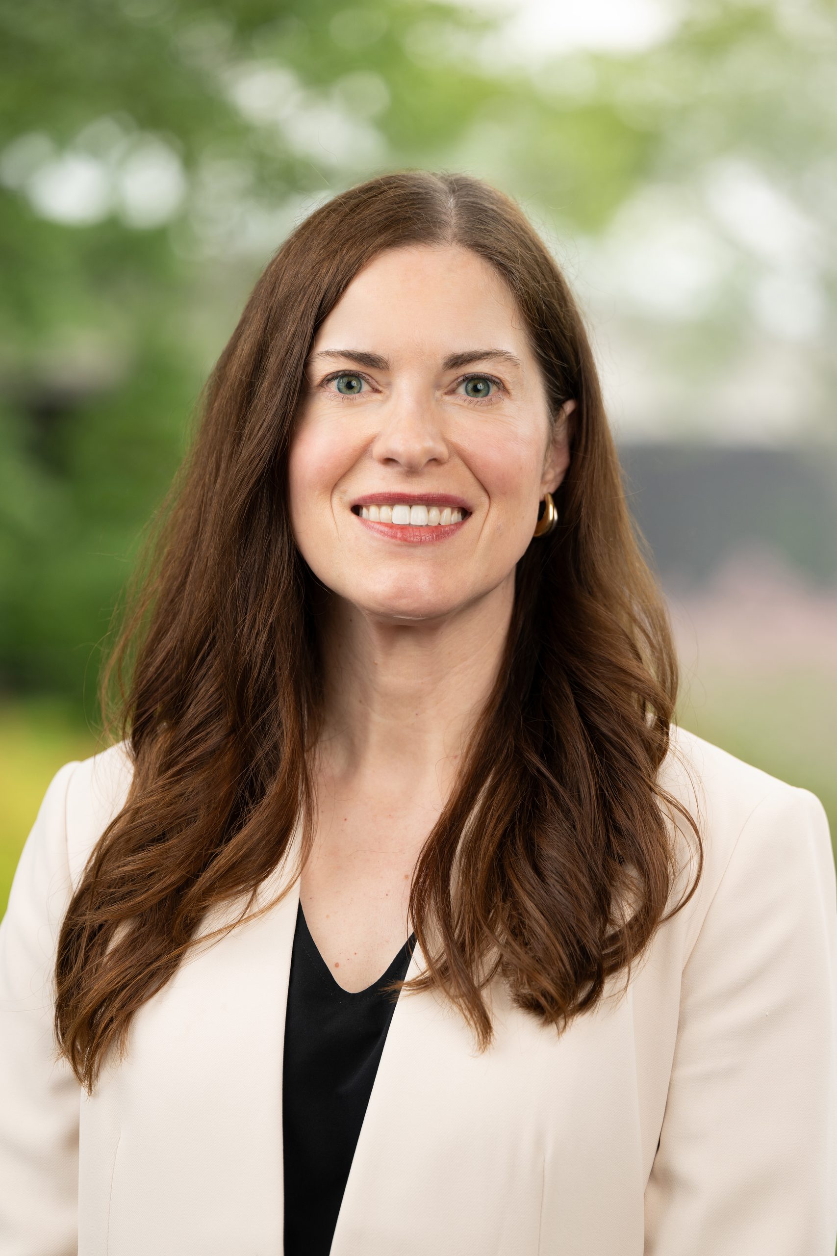 A smiling person with long brown hair, wearing a light-colored blazer over a black top, stands outdoors with a blurred green background.